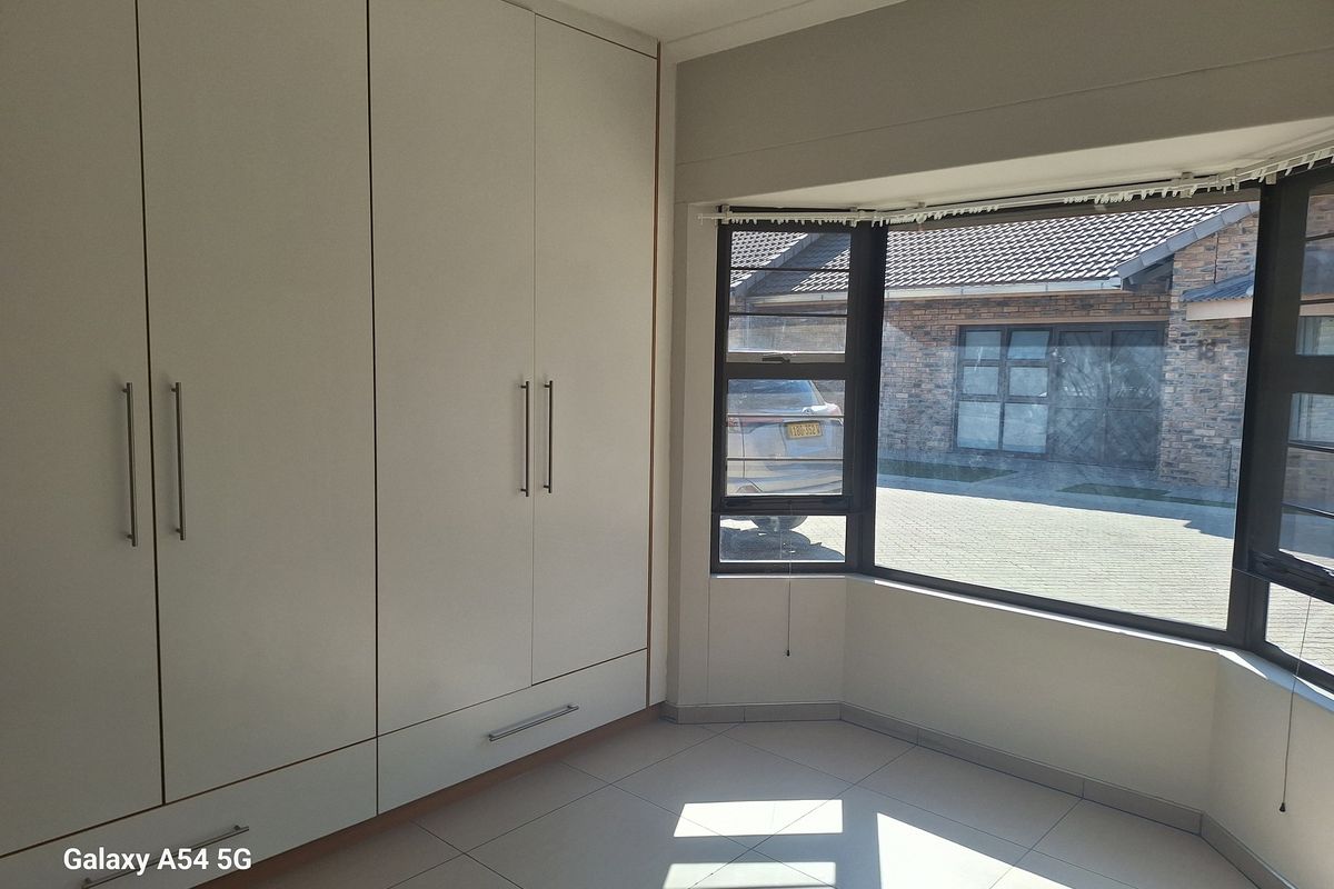 A bright bedroom featuring built-in floor-to-ceiling wardrobes, tiled flooring, and a large bay window overlooking the driveway.