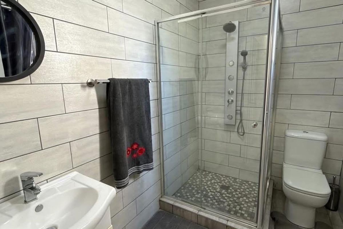 A modern bathroom featuring a glass-enclosed shower with a built-in shower panel, a pedestal sink, and a toilet, finished with light-toned wall tiles.