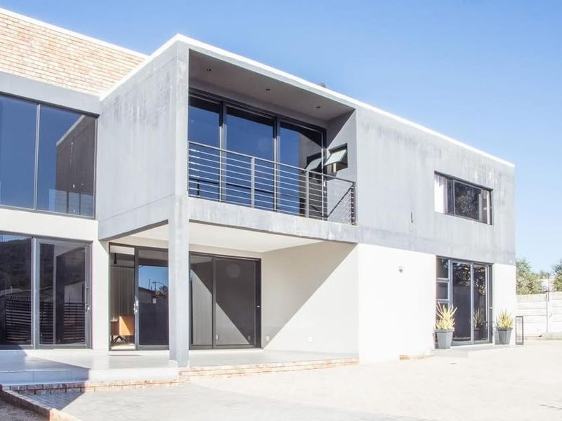 Modern two-story residential exterior featuring a concrete facade, large sliding glass doors, and a balcony with metal railing overlooking a paved driveway.