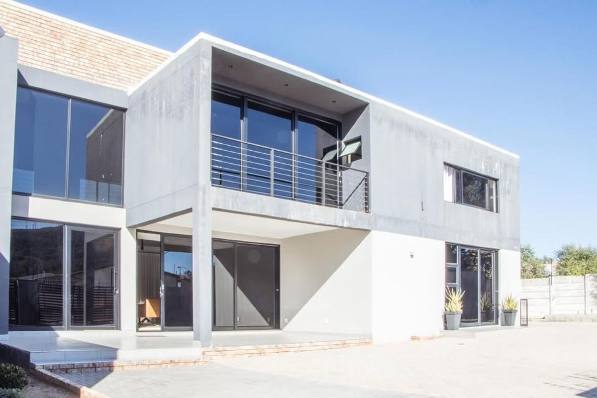 Modern two-story residential exterior featuring a concrete facade, large sliding glass doors, and a balcony with metal railing overlooking a paved driveway.