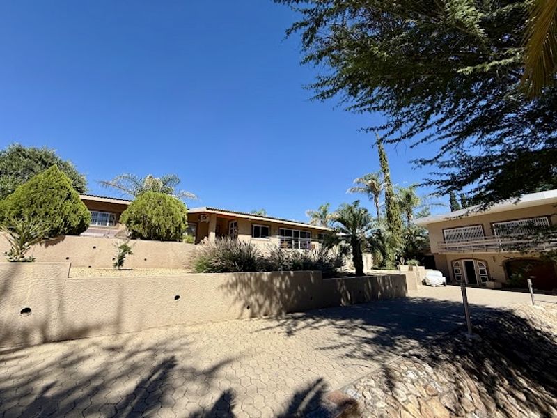 Front view of the property featuring a paved driveway, tiered retaining walls, landscaped garden, and a balcony on the upper level.