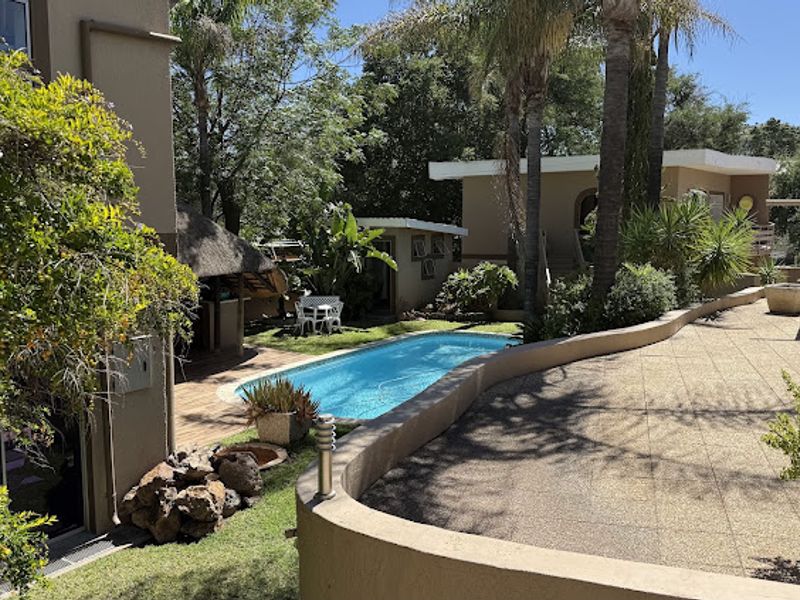 Outdoor entertainment area featuring a swimming pool, paved patio, and a thatched lapa set within a landscaped garden.