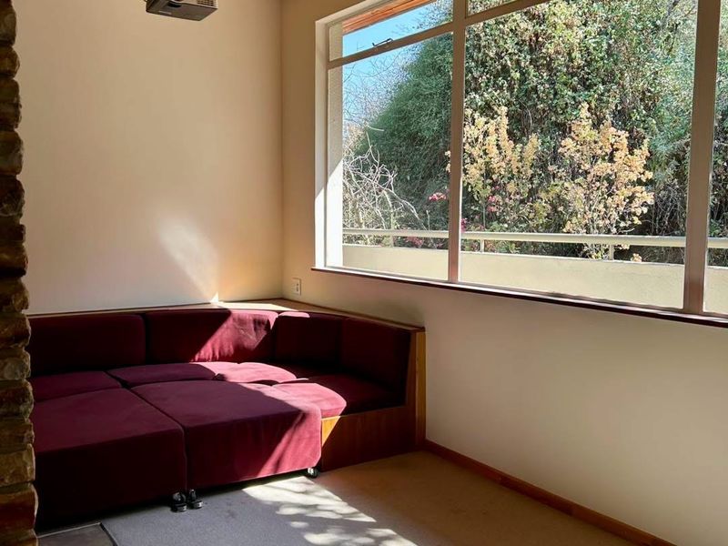 A sunlit living area featuring a timber-clad ceiling, stone accent wall, and a large window overlooking the garden, equipped with a ceiling-mounted projector.
