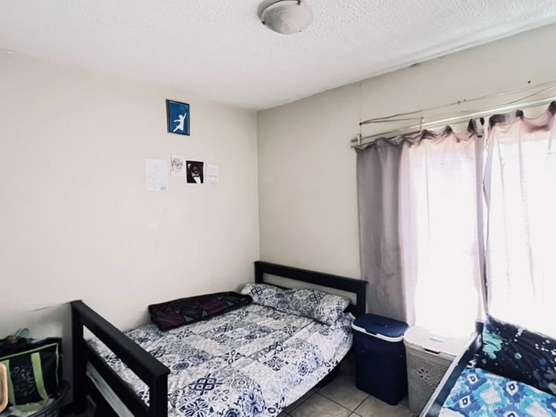 A compact bedroom featuring tiled flooring, a window with curtain rails, and a standard ceiling light fixture.