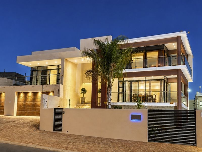 Modern multi-story residence featuring a double garage, expansive glass sliding doors, glass balustrades, and secure boundary wall with integrated lighting.