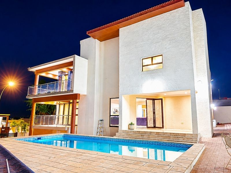 A modern two-story residence featuring a swimming pool, tiled pool deck, and an upper-level balcony with glass balustrades, captured at twilight.
