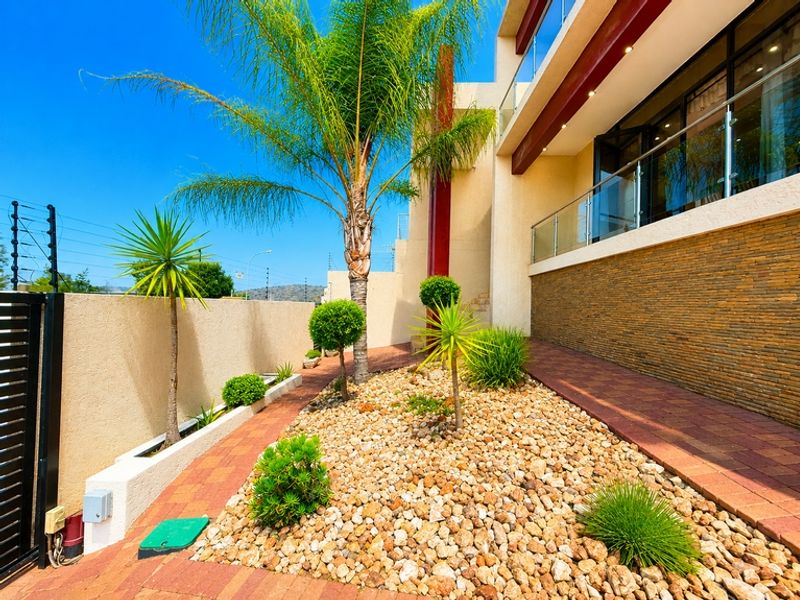 Modern front exterior featuring stone cladding, a paved walkway, and a glass balustrade balcony, complemented by landscaped garden beds.