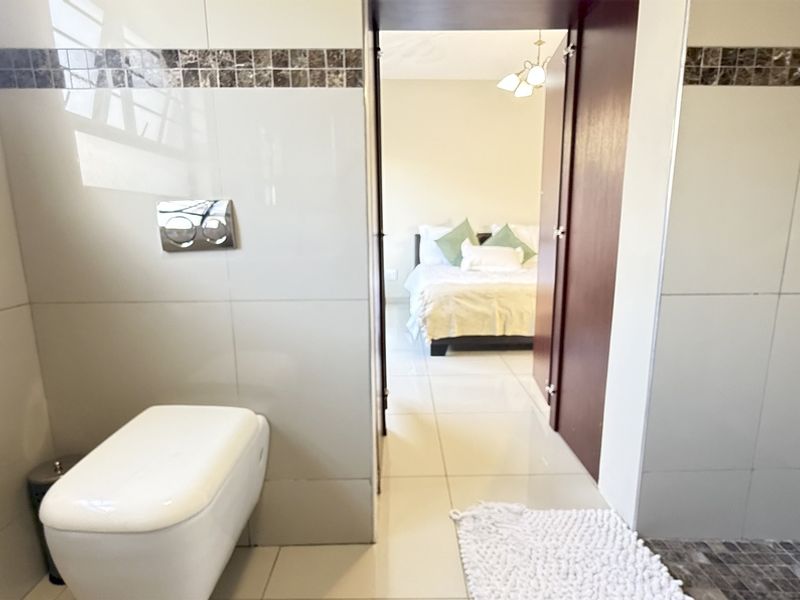 Modern bathroom featuring a wall-mounted toilet with concealed cistern, tiled walls with mosaic accents, and a shower area, leading into an adjacent bedroom.