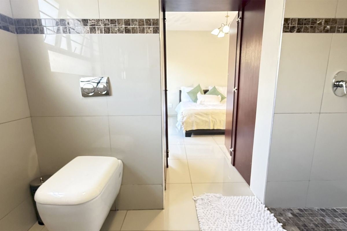 Modern bathroom featuring a wall-mounted toilet with concealed cistern, tiled walls with mosaic accents, and a shower area, leading into an adjacent bedroom.