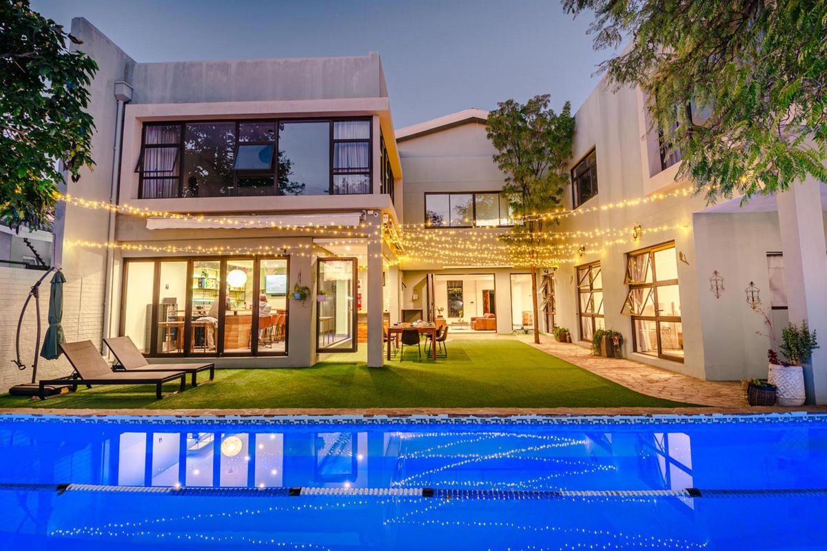 A beautifully illuminated rear exterior view featuring a swimming pool, artificial lawn, and modern multi-story architecture with large glass sliding doors.