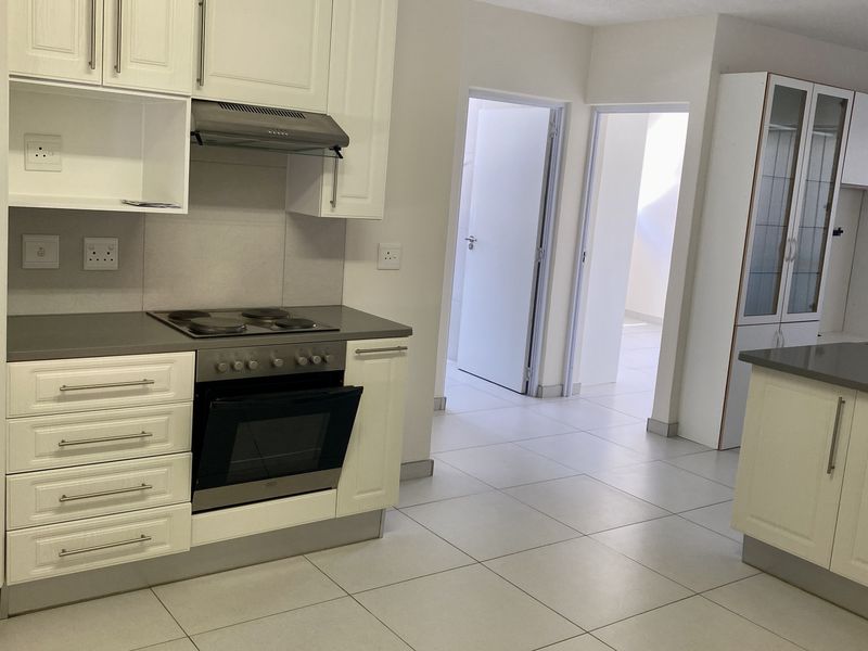 A clean, modern kitchen featuring white cabinetry, stone countertops, integrated oven and hob, and tiled flooring, leading into a hallway.