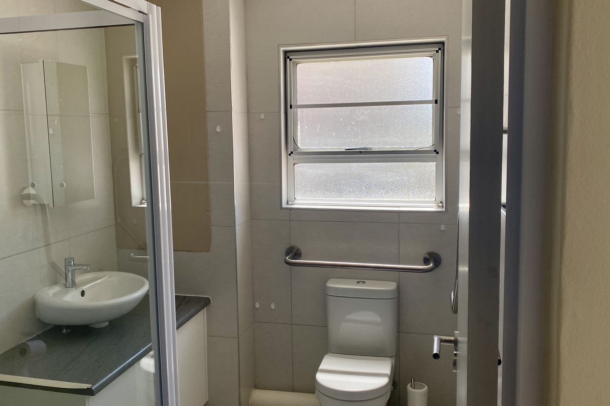 A functional bathroom featuring a vanity with basin, a toilet with a safety grab rail, and a glass-enclosed shower area with tiled walls.
