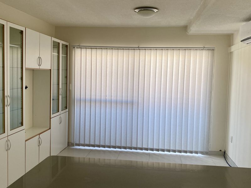 A bright, empty living area featuring built-in storage cabinetry, tiled flooring, vertical blinds, and a wall-mounted air conditioning unit.
