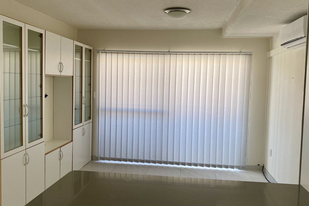 A bright, empty living area featuring built-in storage cabinetry, tiled flooring, vertical blinds, and a wall-mounted air conditioning unit.