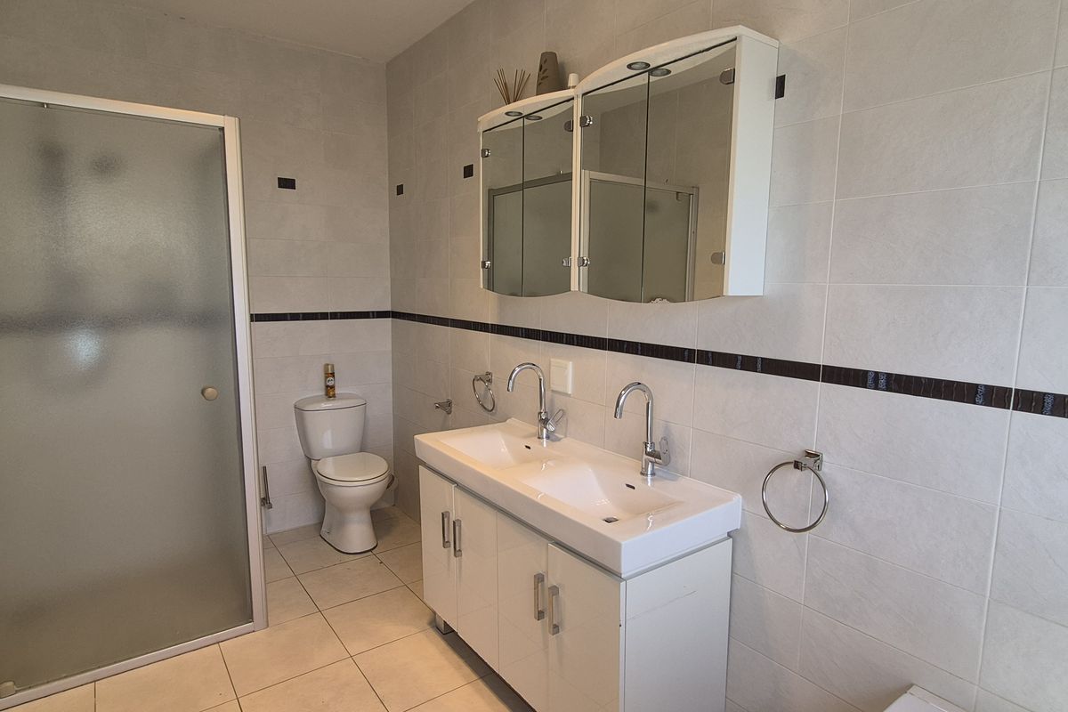 A functional bathroom featuring a double vanity with a mirrored cabinet, a glass-enclosed shower, and a toilet, finished with neutral wall and floor tiles.