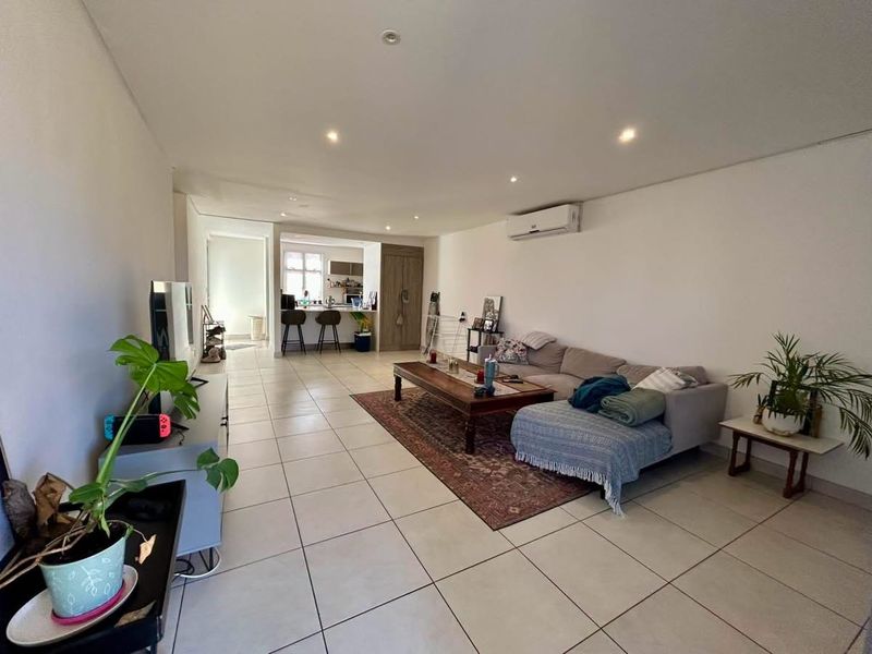 Spacious open-plan living area featuring neutral tiled flooring, recessed lighting, and a wall-mounted air conditioning unit, flowing seamlessly into the kitchen space.