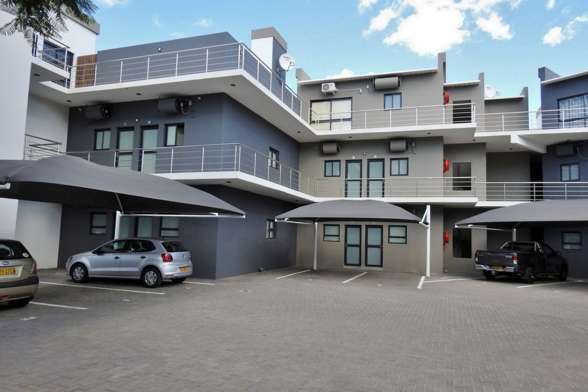 Modern apartment complex exterior featuring paved parking area, shade cloth carports, and multi-level balconies with stainless steel railings.