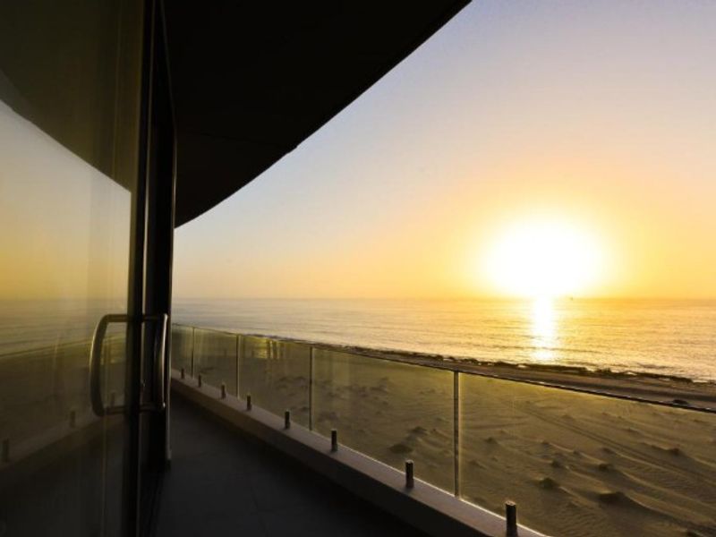A modern balcony featuring a sleek glass balustrade and expansive, unobstructed sunset views over the ocean and coastline.