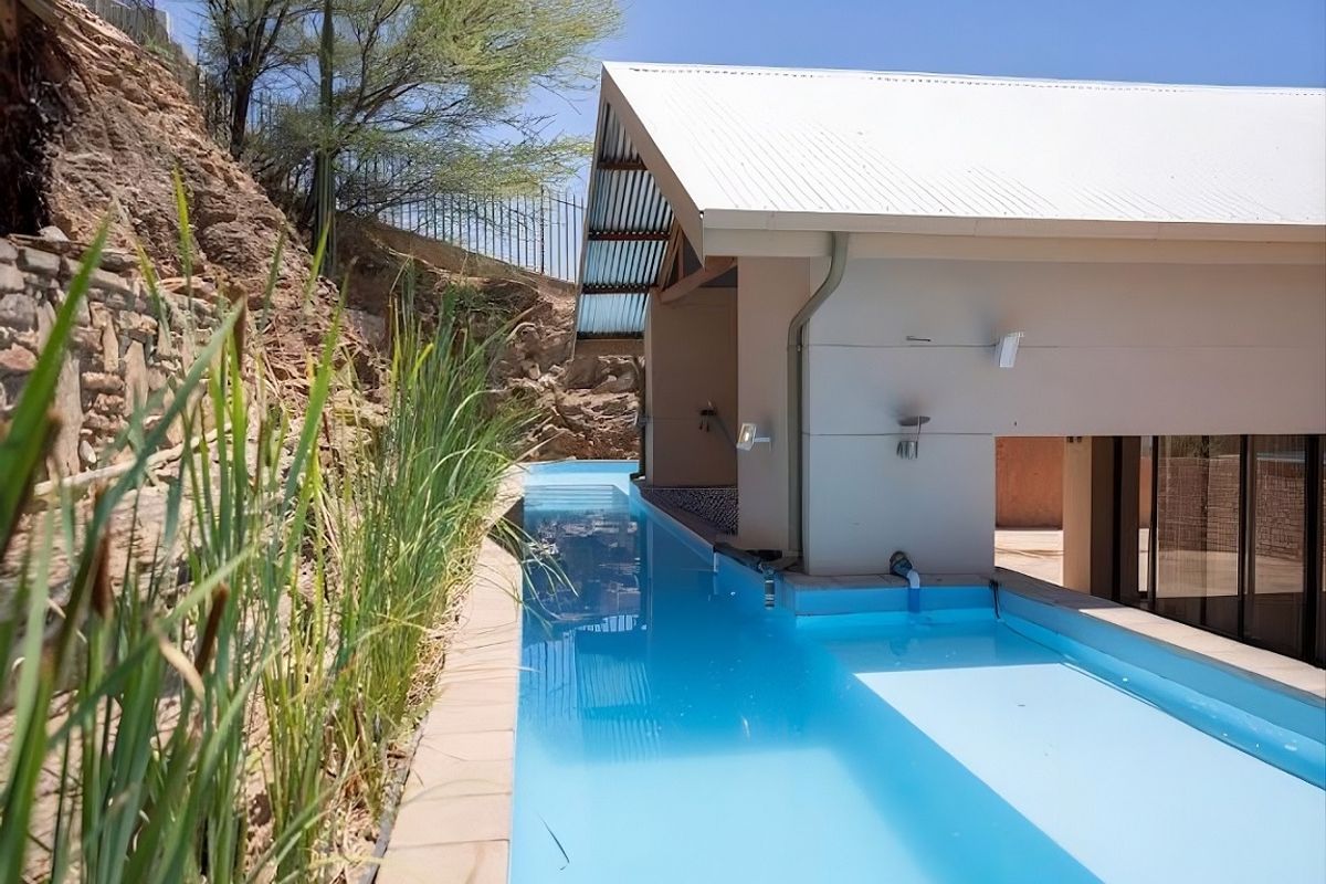 A modern lap pool featuring a covered section with a corrugated metal roof, set against a natural stone retaining wall.