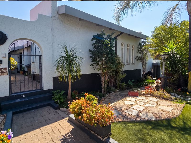 Front entrance area featuring a paved driveway, arched security gate, and landscaped garden with stone pathways and established plantings.