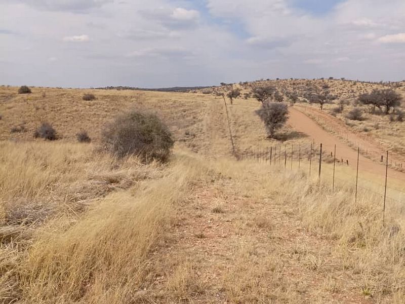 A view of the vacant residential land featuring dry grassland, a dirt access road, and perimeter wire fencing under a cloudy sky.