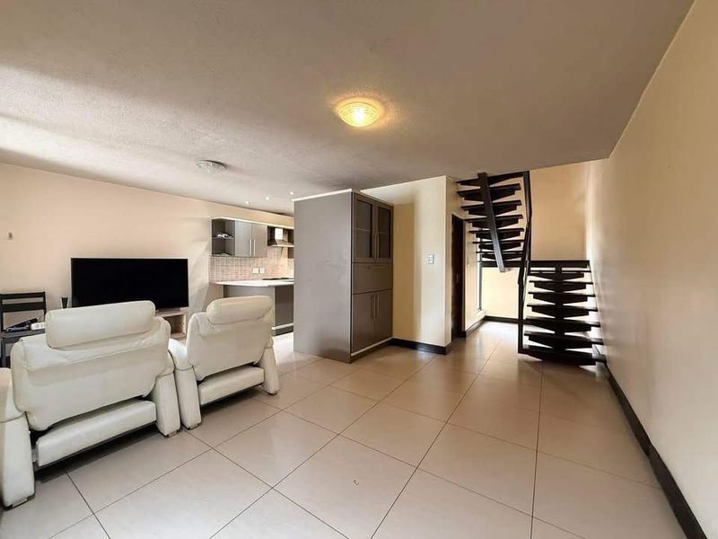 Open-plan living area featuring tiled flooring, a compact kitchen with built-in cabinetry, and a modern staircase leading to the upper level.