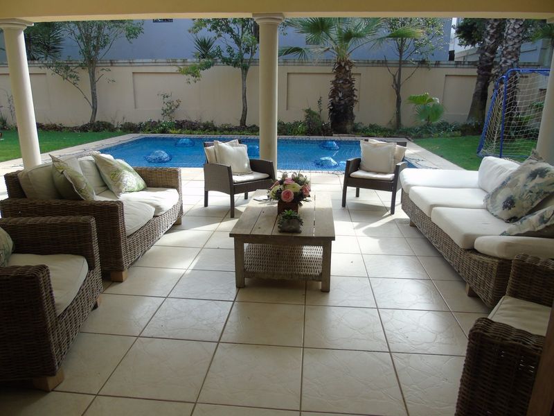 A covered patio area featuring tiled flooring and structural columns, overlooking a private swimming pool and landscaped garden.