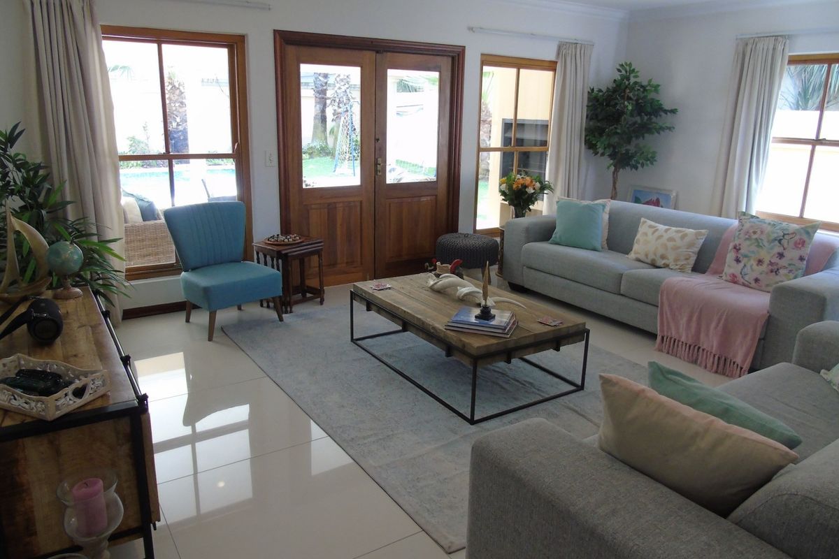 A bright and spacious living room featuring polished tiled floors, large wooden-framed windows, and double doors providing direct access to the outdoor pool area.
