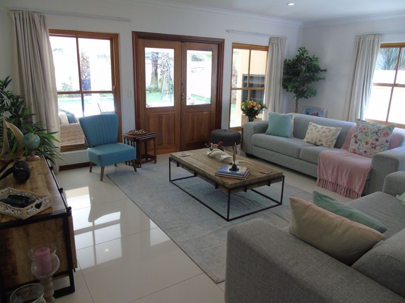 A bright and spacious living room featuring polished tiled floors, large wooden-framed windows, and double doors providing direct access to the outdoor pool area.