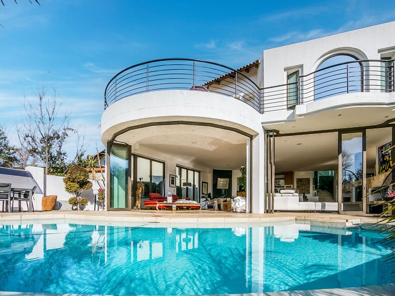 A modern rear exterior view featuring a swimming pool, a spacious covered patio with bi-fold doors, and an upper-level balcony with metal railings.