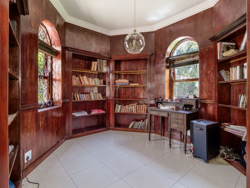 A classic home office featuring rich wood-paneled walls, extensive built-in shelving, arched windows with security grilles, and polished tile flooring.