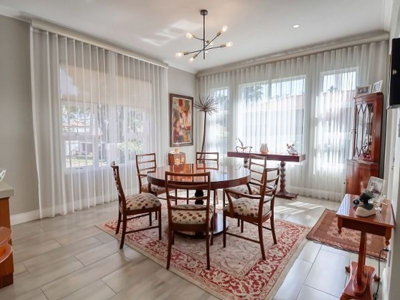A bright and spacious dining room featuring large windows with sheer curtains, modern lighting, and neutral tiled flooring.
