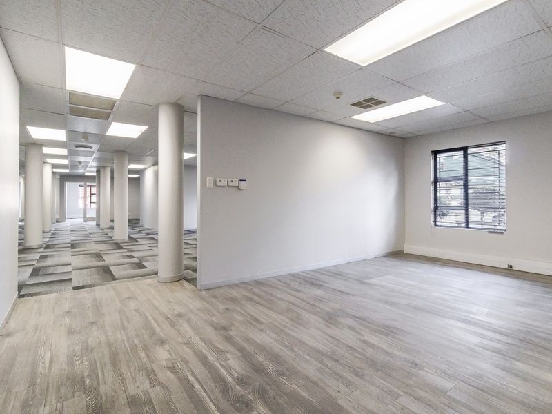 A bright, open-plan office space featuring a mix of laminate and carpet flooring, structural columns, and recessed lighting in a suspended ceiling.