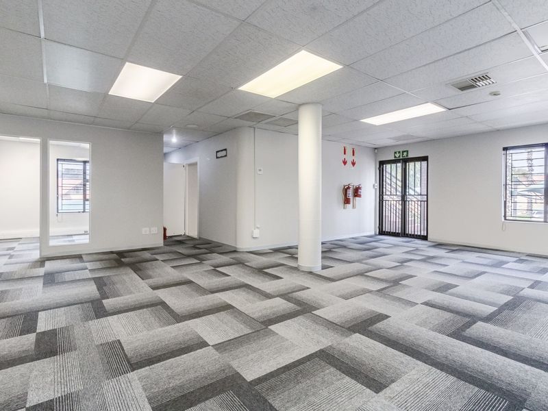A spacious, open-plan office area featuring modern carpet tiles, a suspended ceiling with recessed lighting, and a central structural column.