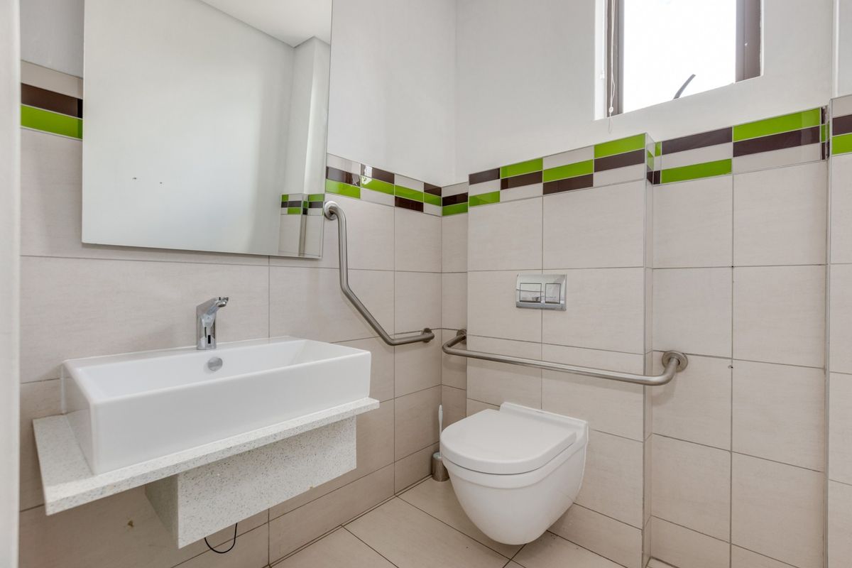 A clean, accessible bathroom featuring a wall-mounted toilet, floating vanity with basin, and chrome grab rails, finished with neutral tiling and decorative accents.