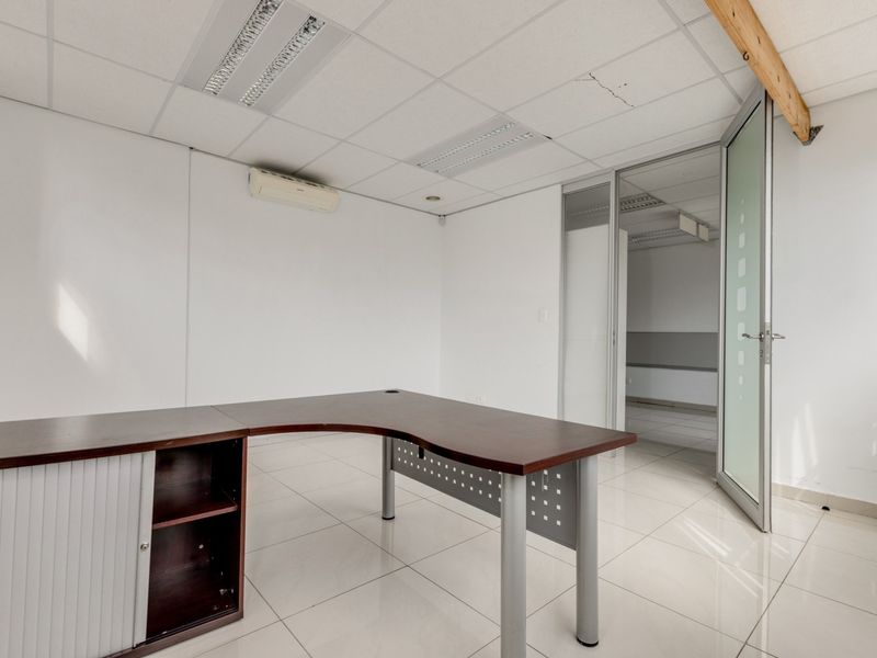A bright, vacant office space featuring tiled flooring, a suspended ceiling with recessed lighting, and a glass partition door leading to an adjacent room.