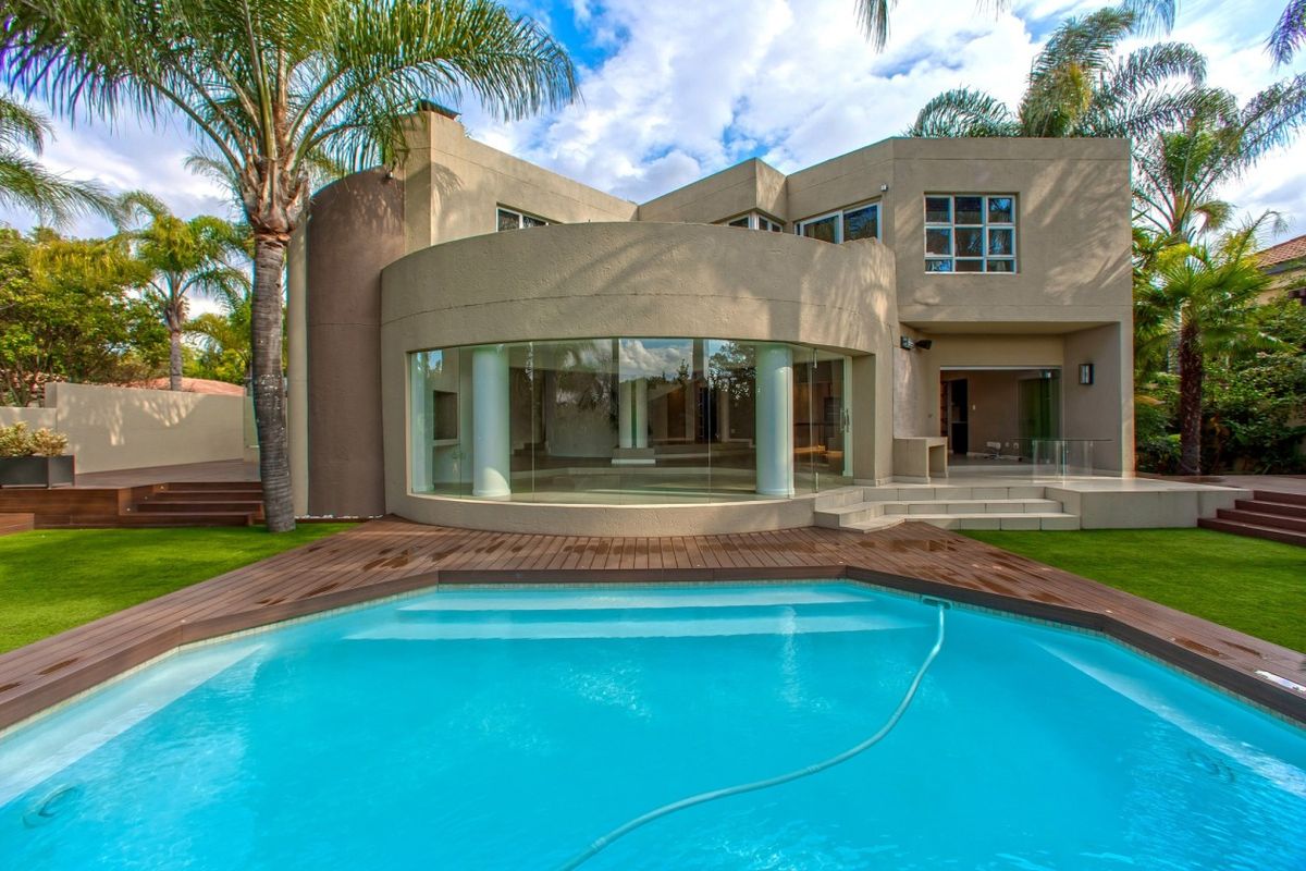 Rear view of a modern residence featuring a swimming pool, timber decking, and a striking curved glass wall with structural columns.