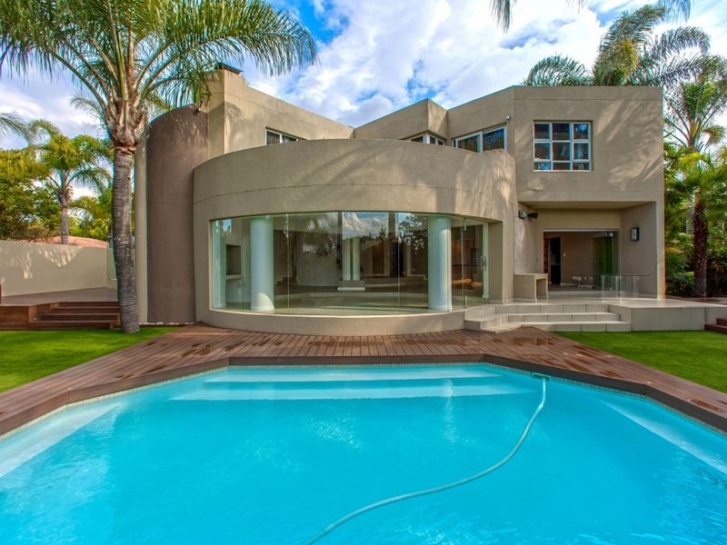 Rear view of a modern residence featuring a swimming pool, timber decking, and a striking curved glass wall with structural columns.