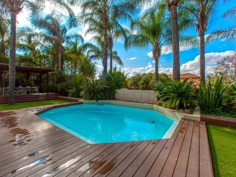 A well-maintained outdoor pool area featuring timber decking, artificial lawn, and a stone water feature, surrounded by mature palm trees.