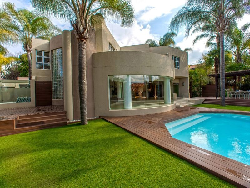 A modern rear exterior view featuring a swimming pool, timber decking, landscaped garden, and a striking curved glass facade with a glass block feature wall.