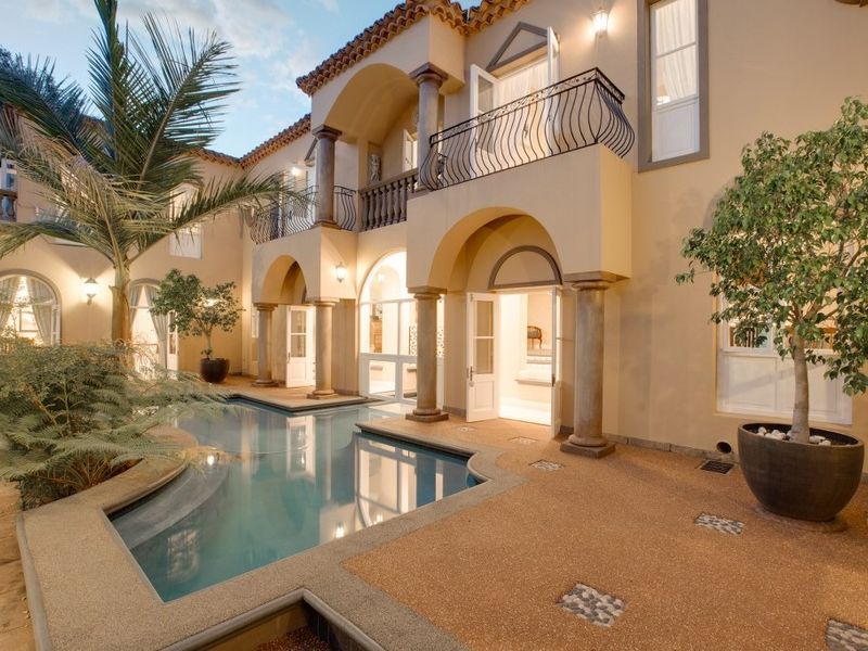 A beautifully maintained Mediterranean-style courtyard featuring a swimming pool, covered patio, and upper-level balcony with elegant stone columns and wrought iron detailing.