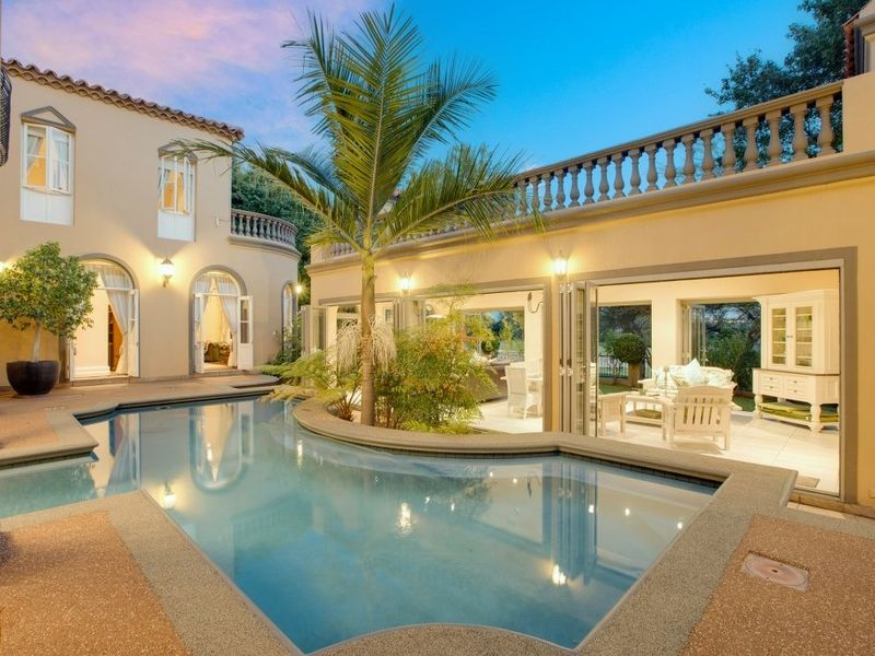 A beautifully maintained swimming pool area featuring a curved pool design, stone-tiled patio, and seamless indoor-outdoor flow through large bi-fold doors.