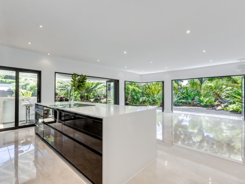 Spacious open-plan kitchen area featuring a modern island with built-in appliances, sleek tiled flooring, and expansive bi-fold doors opening to a lush garden.