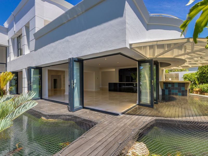 Rear exterior view featuring a modern home with bi-fold doors opening onto a timber deck, pond, and a built-in outdoor bar area.