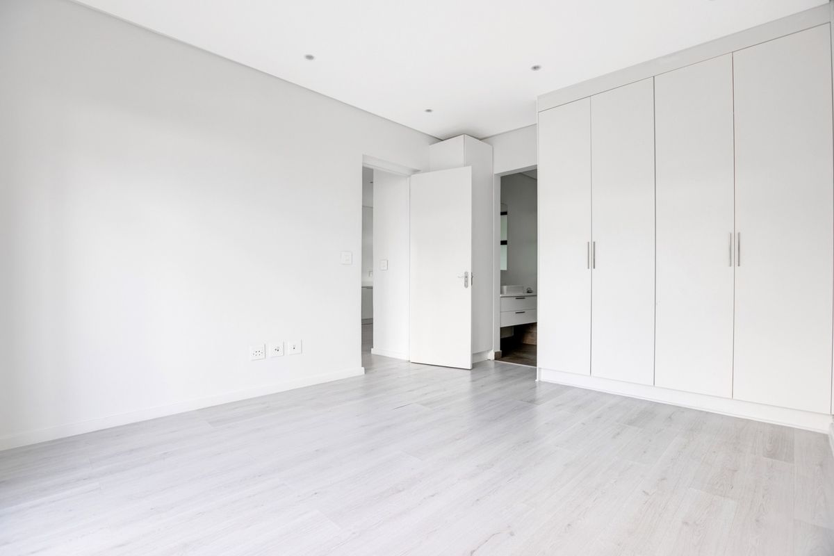 A bright, empty bedroom featuring modern laminate flooring, built-in cupboards, and recessed downlighting, with an adjacent en-suite bathroom visible.