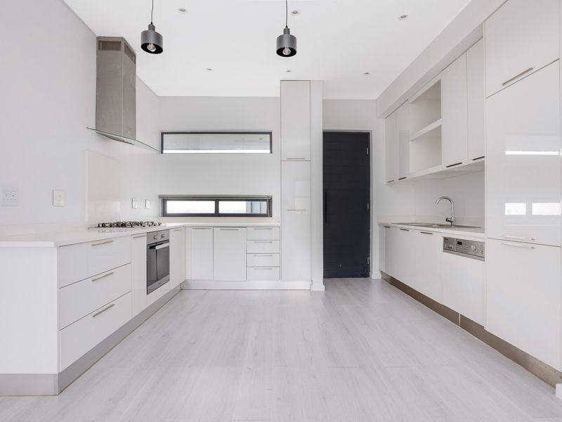 A modern, minimalist kitchen featuring white cabinetry, integrated appliances, a gas hob, and sleek laminate flooring in a bright, open-plan layout.