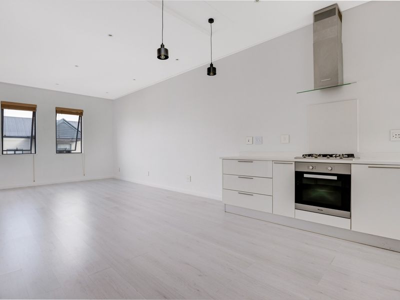 A bright, modern open-plan living and kitchen area featuring light laminate flooring, sleek white cabinetry, built-in appliances, and contemporary pendant lighting.