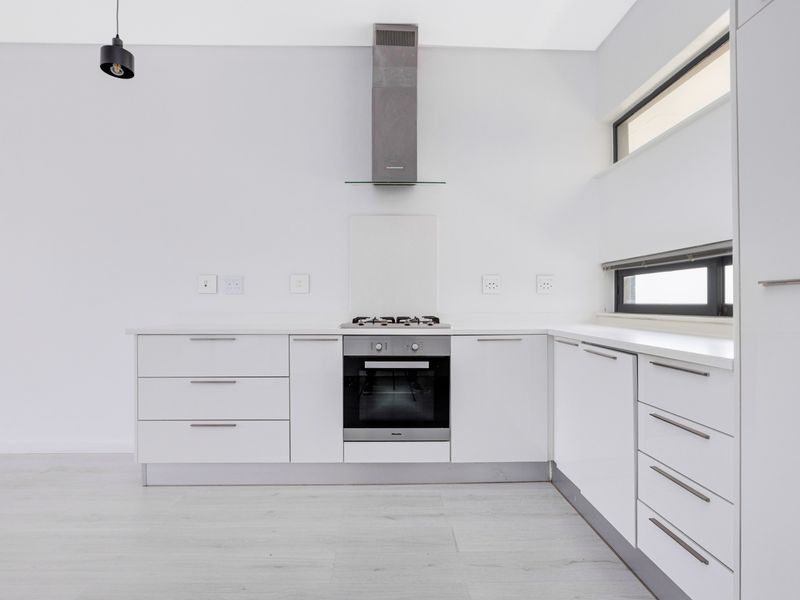 Modern, minimalist kitchen featuring white cabinetry, stone countertops, integrated oven, gas hob, and extractor fan in a clean, bright, and empty space.