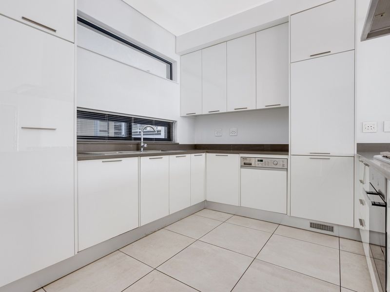 A modern, minimalist kitchen featuring white high-gloss cabinetry, stone countertops, integrated appliances, and neutral floor tiles in a clean, well-lit space.