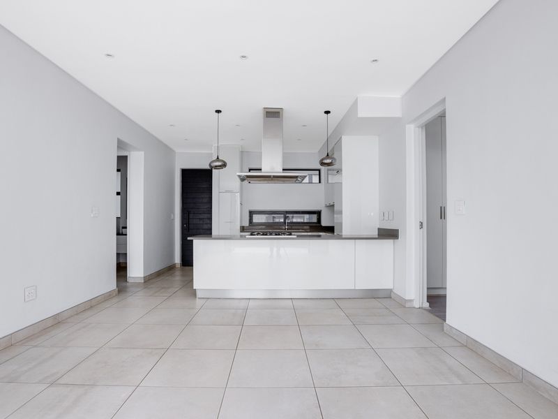 A modern, open-plan kitchen and living area featuring sleek white cabinetry, a central island with a gas hob, and neutral tiled flooring throughout.